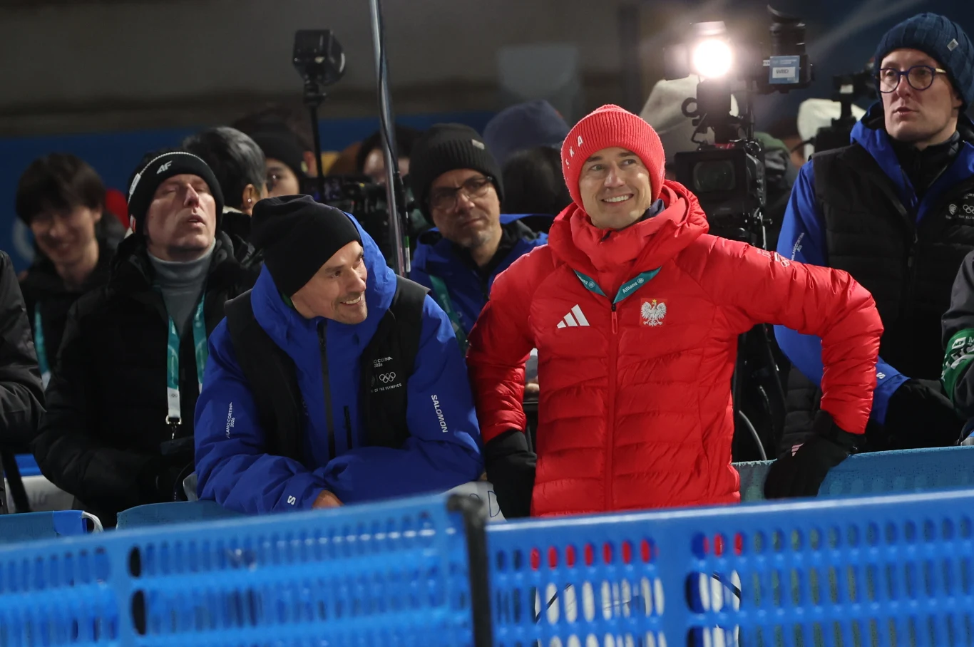 Piotr Żyła i Kamil Stoch Osoba w czerwonym stroju sportowym z biało-czerwonym godłem Polski na piersi stoi wśród grupy ludzi ubranych w zimowe kurtki, niektórzy z nich mają sprzęt fotograficzny, za niebieskimi barierkami ustawionymi na tle areny sportowej.