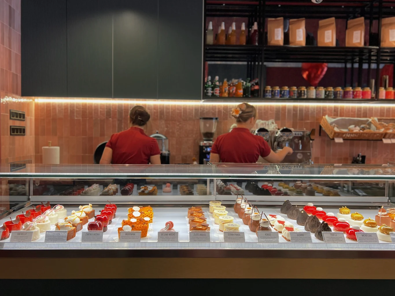 Witryna pełna różnorodnych, kolorowych deserów i ciast, za którą pracują dwie osoby w bordowych uniformach, przygotowując zamówienia. Na półkach w tle ustawione są słoiki, opakowania oraz pieczywo.