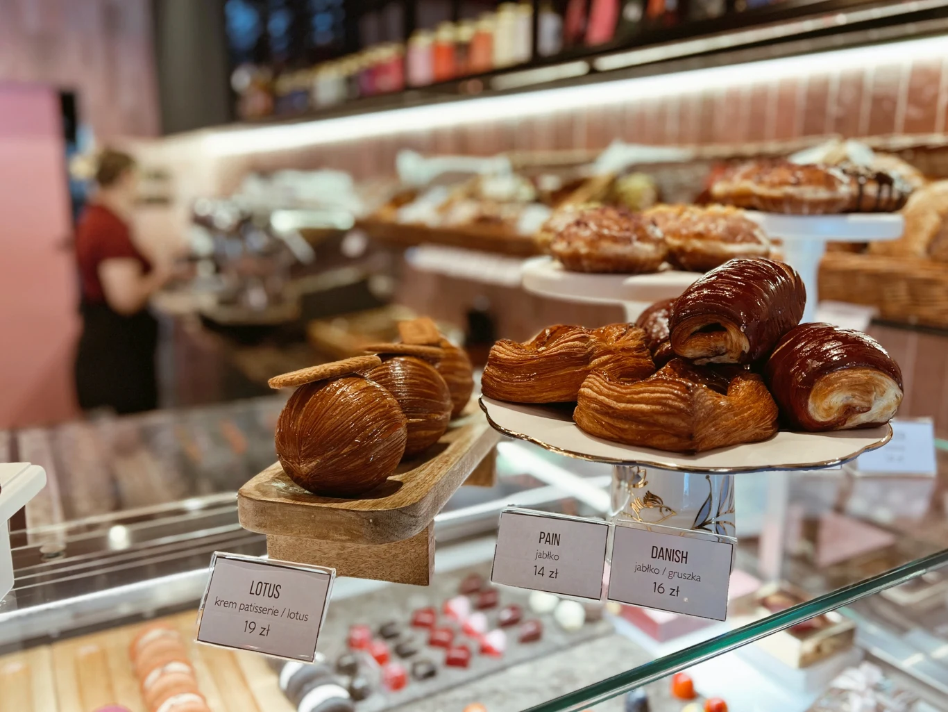 Witryna z różnorodnymi wypiekami, wśród których znajdują się maślane bułki, rogaliki oraz inne słodkie drożdżówki, z widocznymi cenami i opisami, w tle rozmyte wnętrze kawiarni.