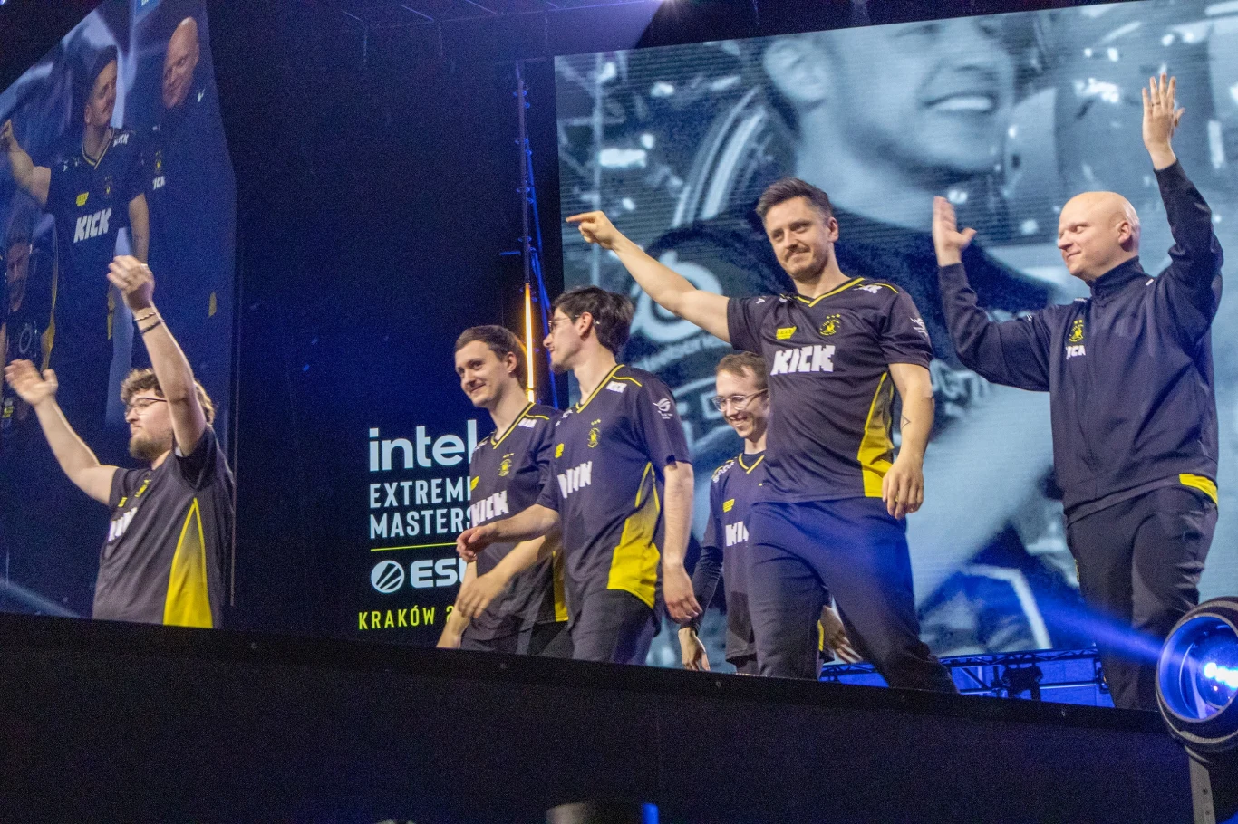 Grupa zawodników drużyny e-sportowej ubrana w firmowe koszulki, stojących na scenie i machających do publiczności, za nimi duży ekran z wyświetlonym powiększonym obrazem wydarzenia.