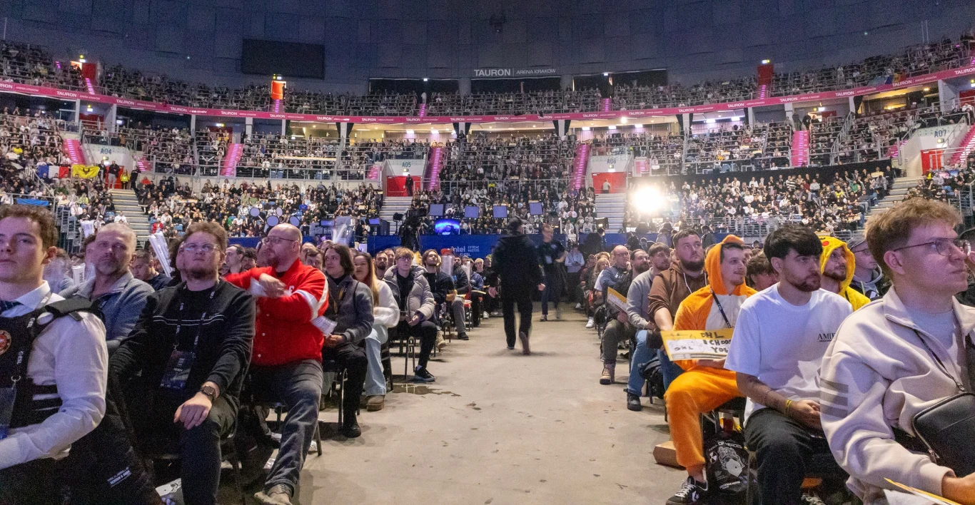 Tłum ludzi siedzących na trybunach i krzesłach w dużej hali widowiskowej, oświetlonej podczas dużego wydarzenia. Wiele osób patrzy w stronę sceny, część ubrana bardziej swobodnie, niektórzy w kostiumach. Atmosfera skupienia i oczekiwania.