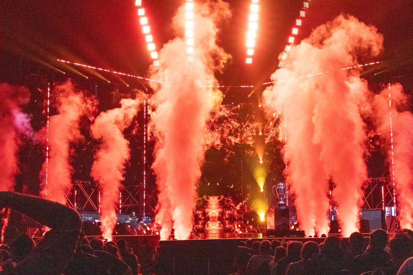 Widownia obserwuje scenę koncertową z intensywnymi efektami świetlnymi oraz wyrzutniami dymu i kolorowych świateł; dominują ciepłe barwy oraz dynamiczny klimat widowiska.