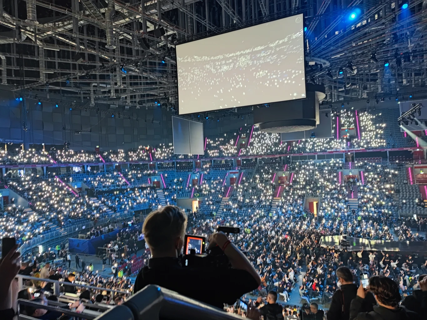 Pełna publiczności hala koncertowa, tysiące widzów świecących latarkami telefonów, scena centralna i duży ekran LED pod sufitem, widoczne reflektory i nowoczesna infrastruktura oświetleniowa.