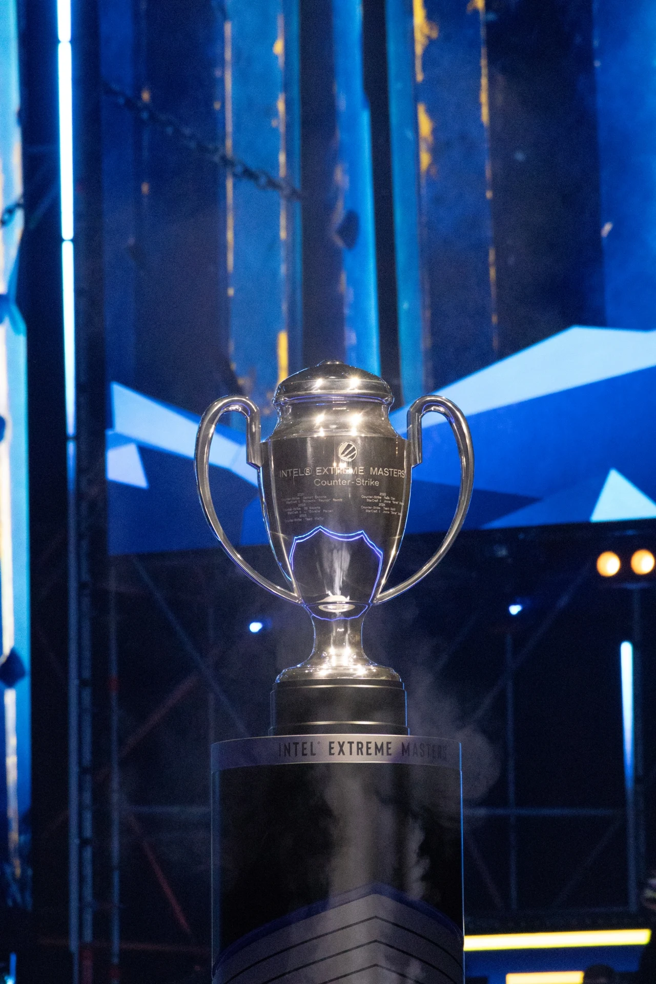 Srebrny puchar z wygrawerowanym napisem Intel Extreme Masters ustawiony na czarnym postumencie, podświetlony niebieskim światłem na tle elementów scenograficznych o industrialnym charakterze.