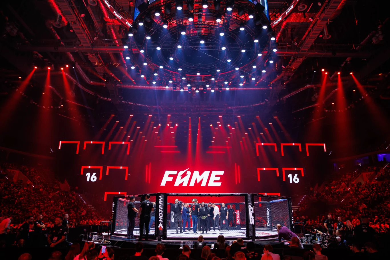 FAME MMA Tłum zgromadzony wokół oktagonu na wielkiej gali sportów walki, efektowne oświetlenie sceniczne, liczni uczestnicy oraz napis 'FAME' w centrum uwagi. Widownia wypełnia arenę, panuje pełna energii atmosfera.
