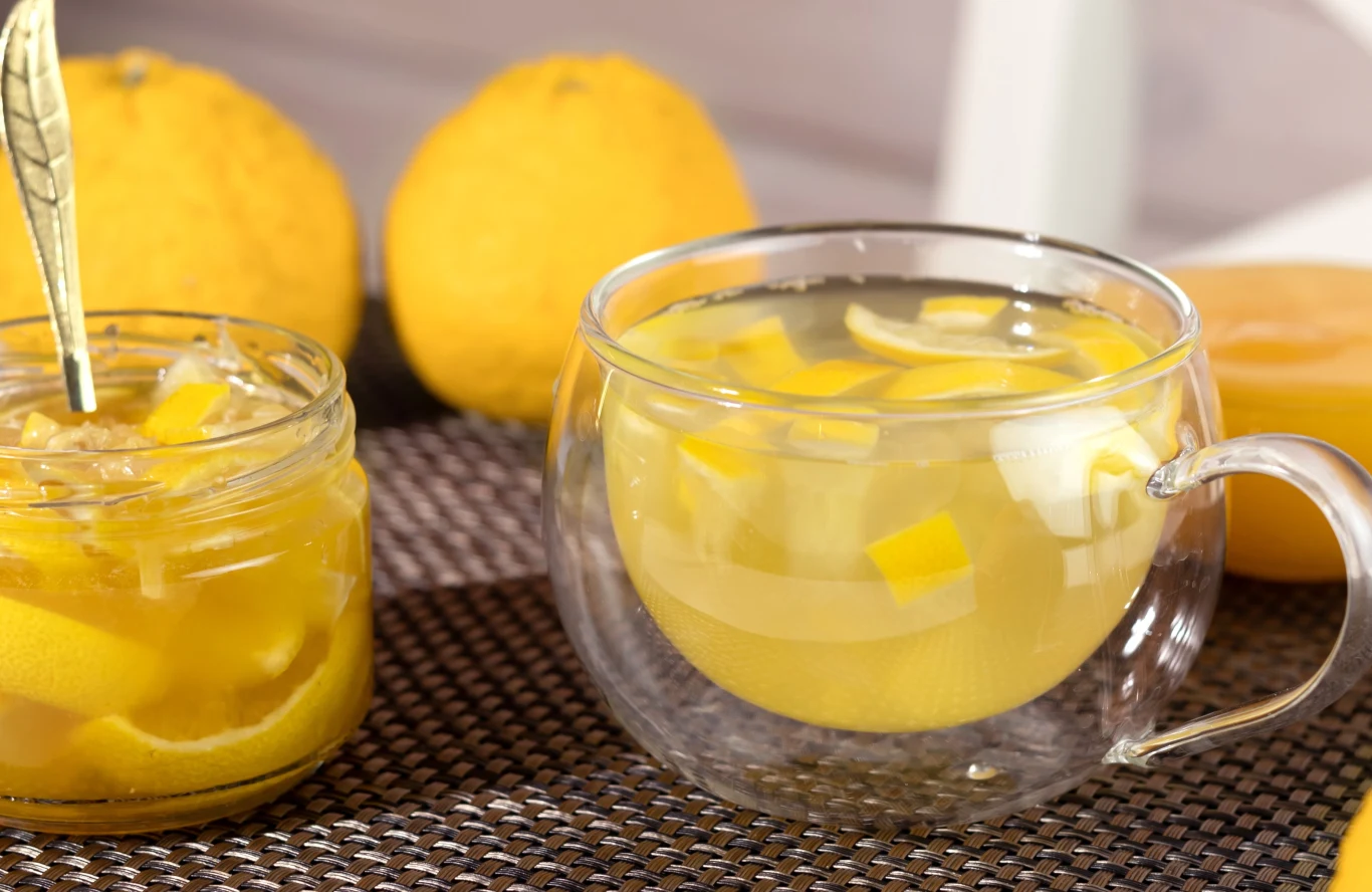 Popularny napar z yuzu to świetnie rozgrzewające remedium na przeziębienie Szklanka z napojem cytrynowym pełnym kawałków świeżej cytryny, obok słoik z plasterkami cytryny i łyżeczką, w tle dwie całe cytryny, wszystko ustawione na plecionej podkładce.