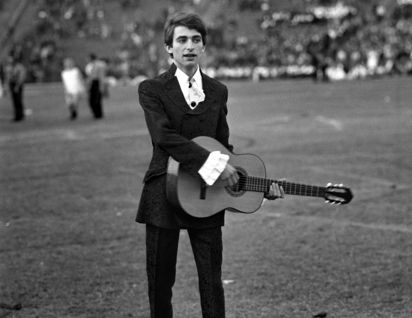 Seweryn Krajewski pod koniec lat 60. Młody mężczyzna w eleganckim garniturze trzyma gitarę akustyczną na stadionie, w tle rozmyte sylwetki ludzi oraz trybuny wypełnione publicznością.