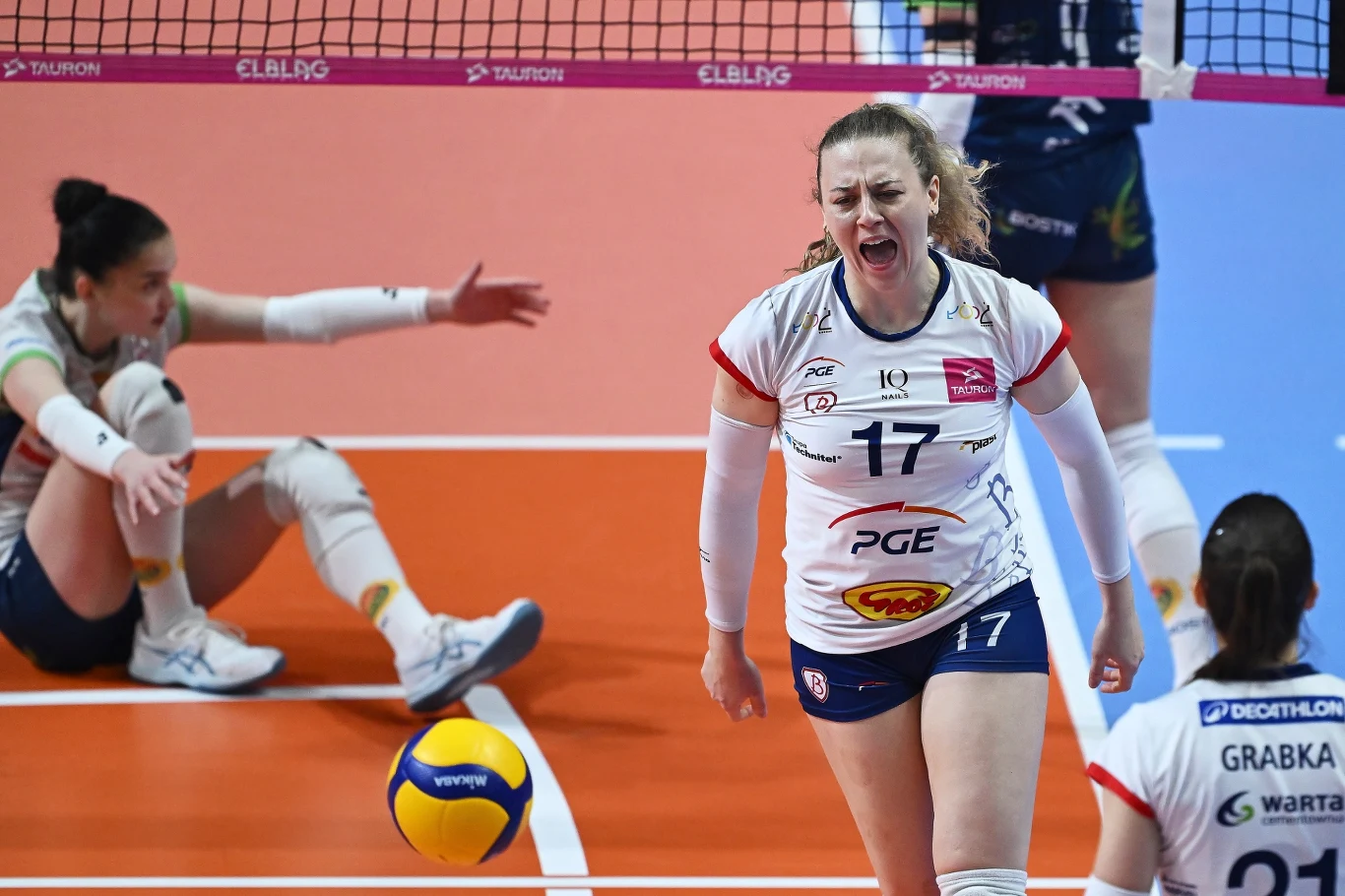 Siatkarka w białym stroju wyraża silne emocje po akcji podczas meczu, w tle inna zawodniczka siedzi na parkiecie, obok leży piłka siatkowa. Wszystko odbywa się na hali sportowej.