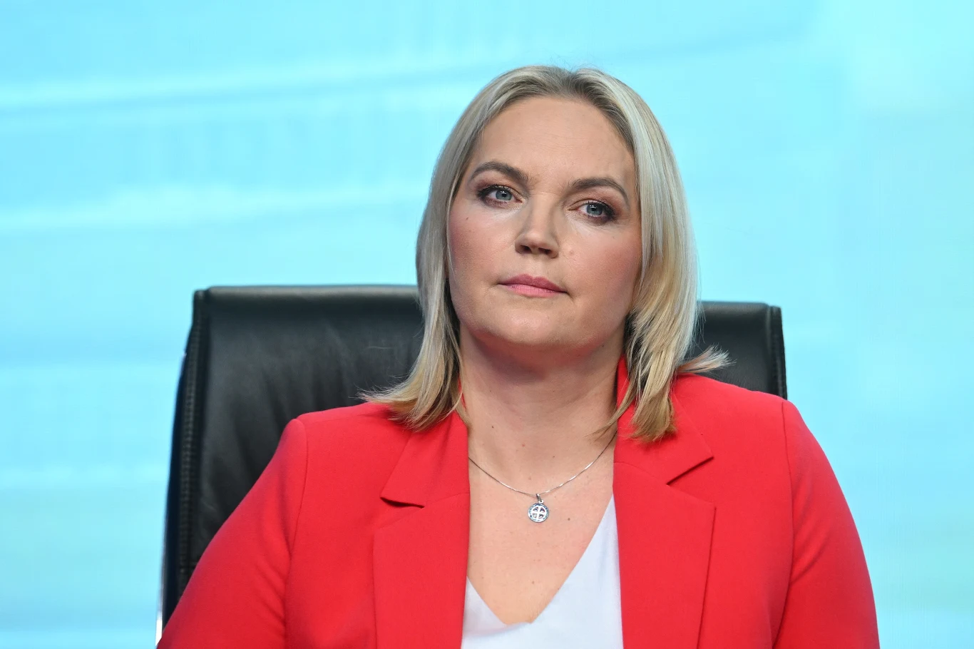 Kobieta o blond włosach w czerwonej marynarce, siedząca na czarnym krześle, na jasnym niebieskim tle, z poważnym wyrazem twarzy.