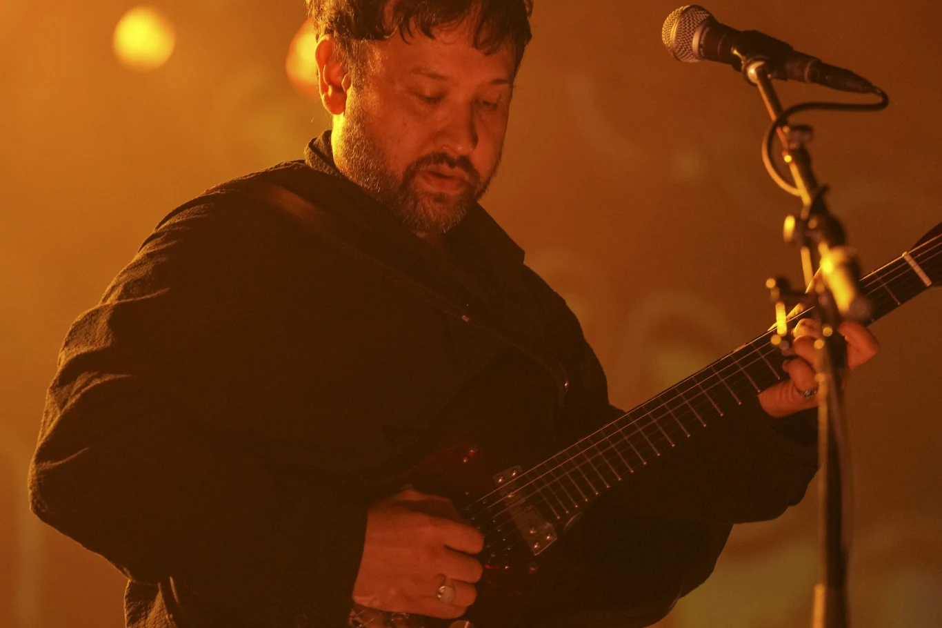 Ruban Nielson Mężczyzna grający na gitarze elektrycznej na tle sceny koncertowej, delikatnie oświetlony ciepłym, pomarańczowym światłem, z mikrofonem umieszczonym blisko jego ust.