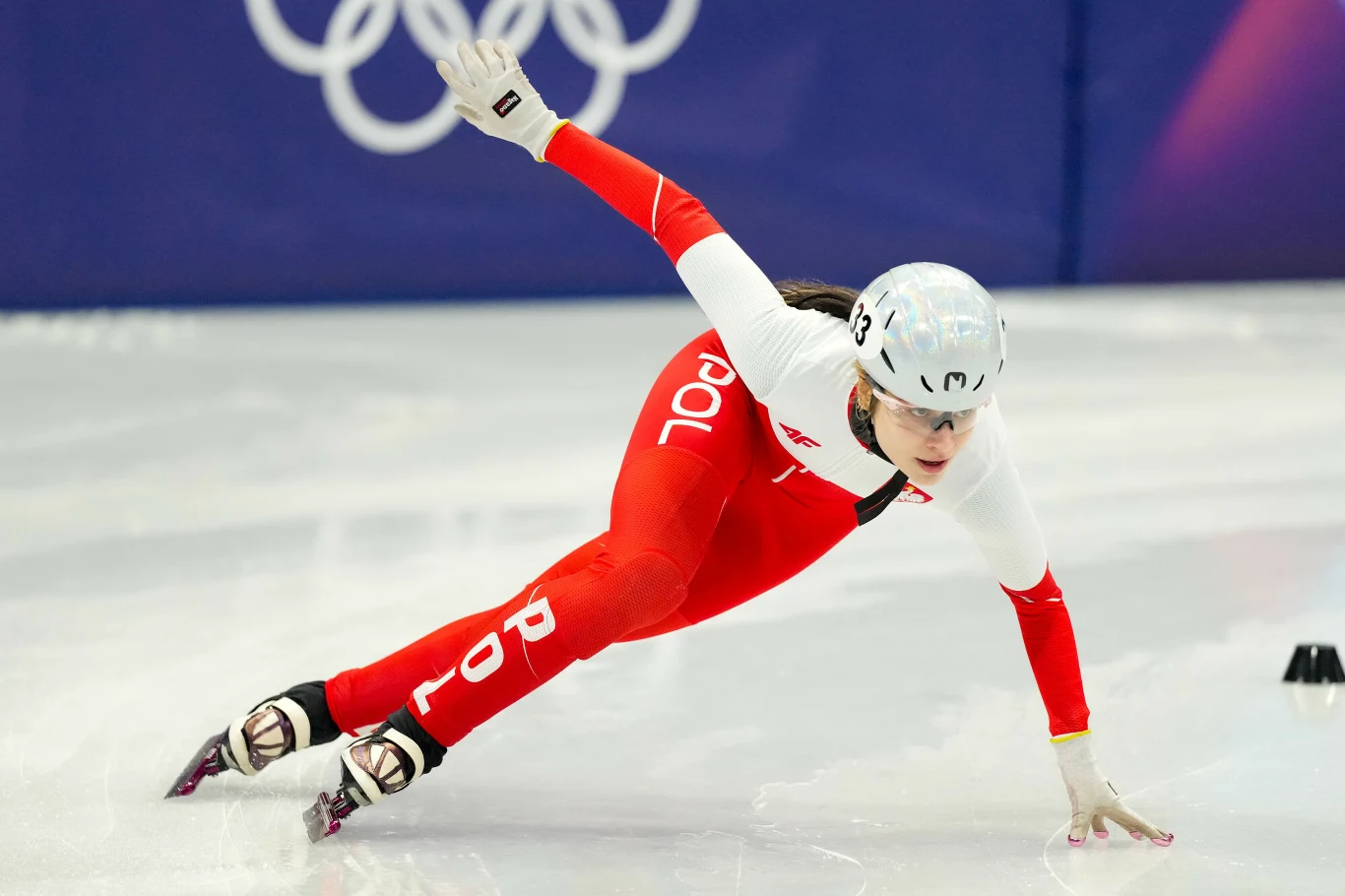 Gabriela Topolska niosła polską flagę wraz z Władimirem Semirunnijem w trakcie ceremonii zamknięcia ZIO 2026 Łyżwiarka szybka w biało-czerwonym stroju z napisem 'POL' w dynamicznej pozie podczas wyścigu na lodowej tafli, w tle widoczne symbole olimpijskie.