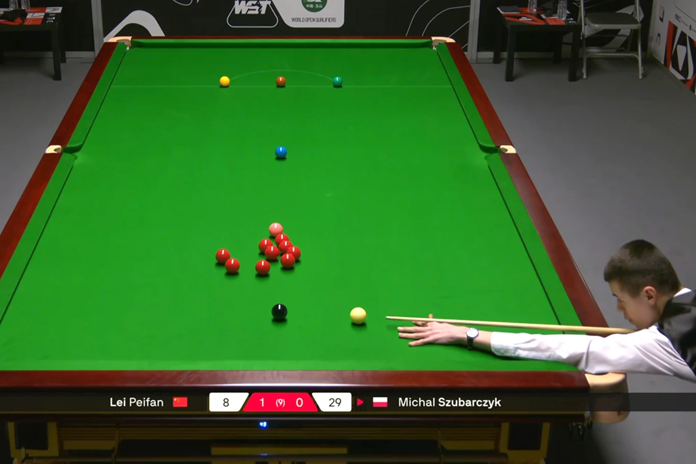 Zawodnik wykonuje uderzenie przy stole snookerowym, na którym rozłożone są kolorowe bile, obok tablica wyników z imionami graczy i aktualnym stanem punktowym.