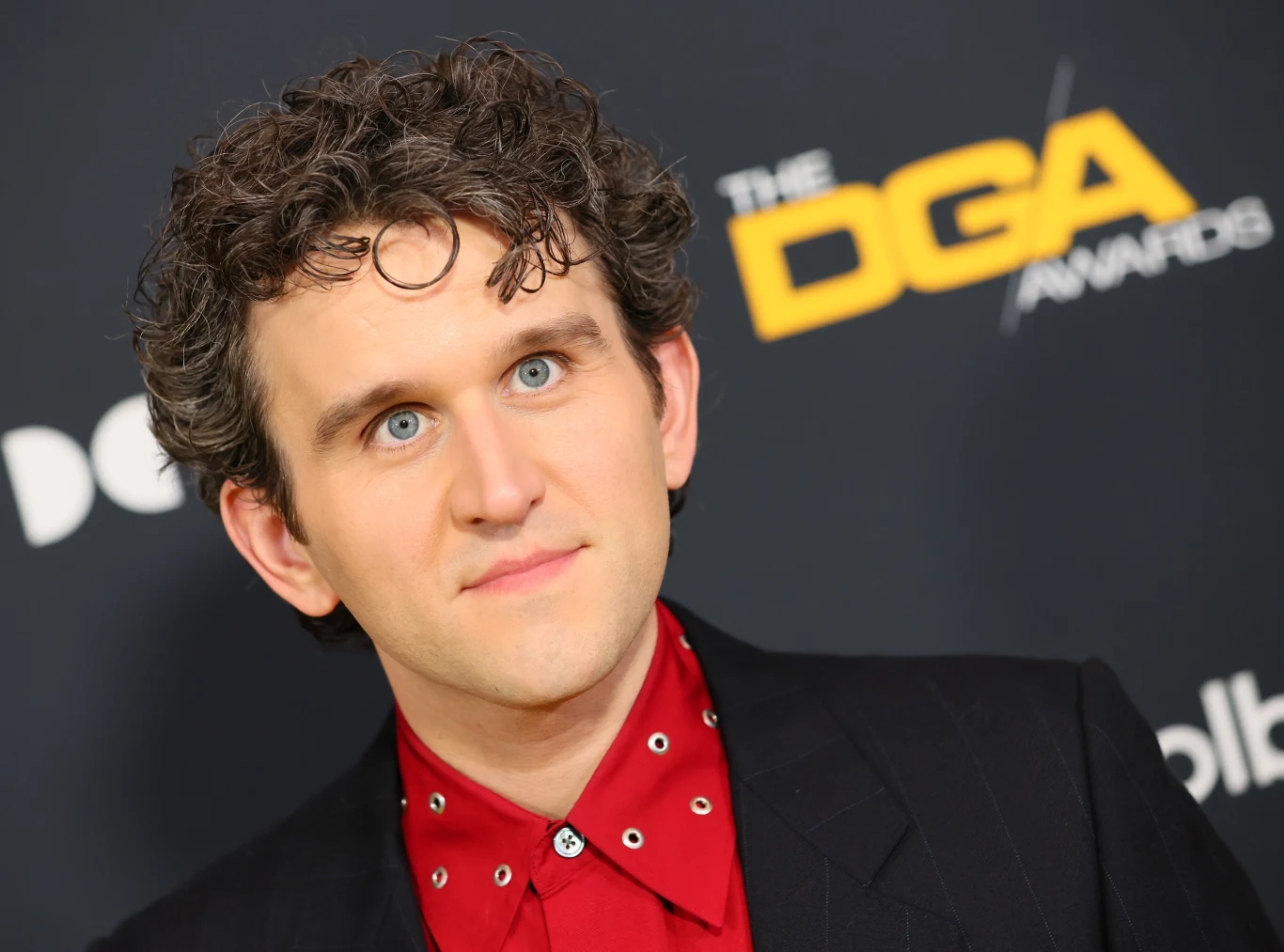 Harry Melling Mężczyzna o kręconych brązowych włosach, jasnej cerze i niebieskich oczach, ubrany w czarną marynarkę i czerwoną koszulę z ozdobnymi elementami, stoi na tle ścianki z logotypem DGA AWARDS.