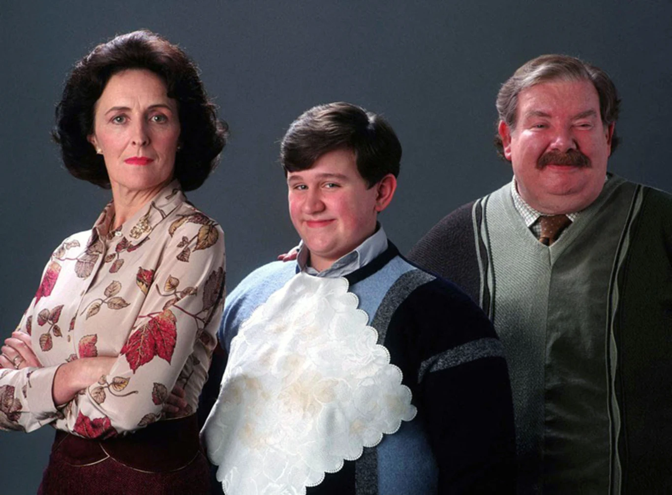Richard Griffiths, Fiona Shaw, Harry Melling na planie "Harry'ego Pottera" Trzy osoby stojące obok siebie na tle ciemnego, neutralnego tła, po lewej kobieta z krótkimi, ciemnymi włosami w eleganckiej bluzce, po środku młody chłopak w swetrze z dużą białą serwetką, po prawej mężczyzna z wąsem w zielonym swetrze, wszyscy patrzą...