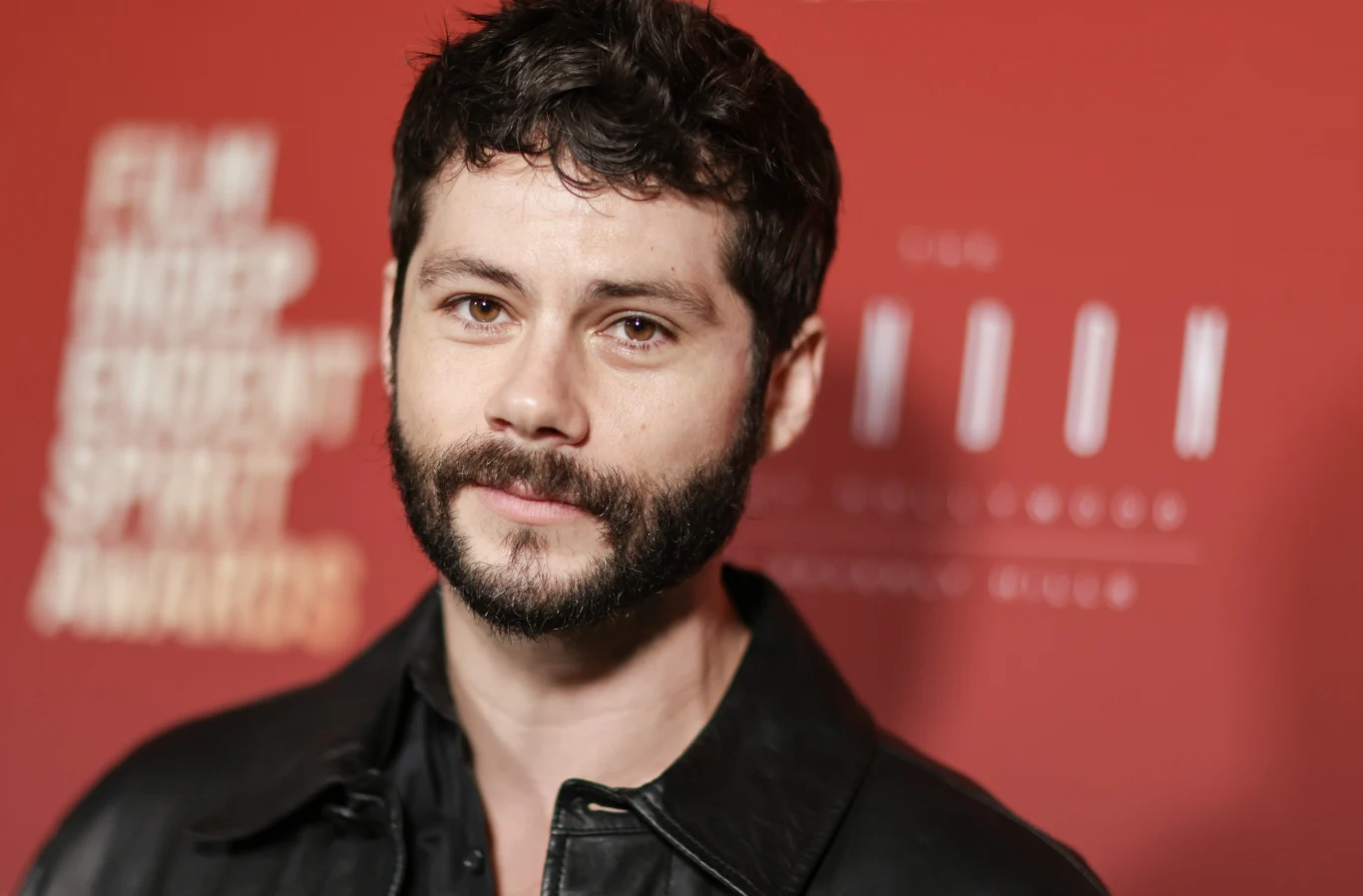 Dylan O'Brien Mężczyzna z krótkimi, ciemnymi włosami i brodą, ubrany w czarną kurtkę, stoi na tle czerwonej ścianki z niewyraźnym napisem wydarzenia.