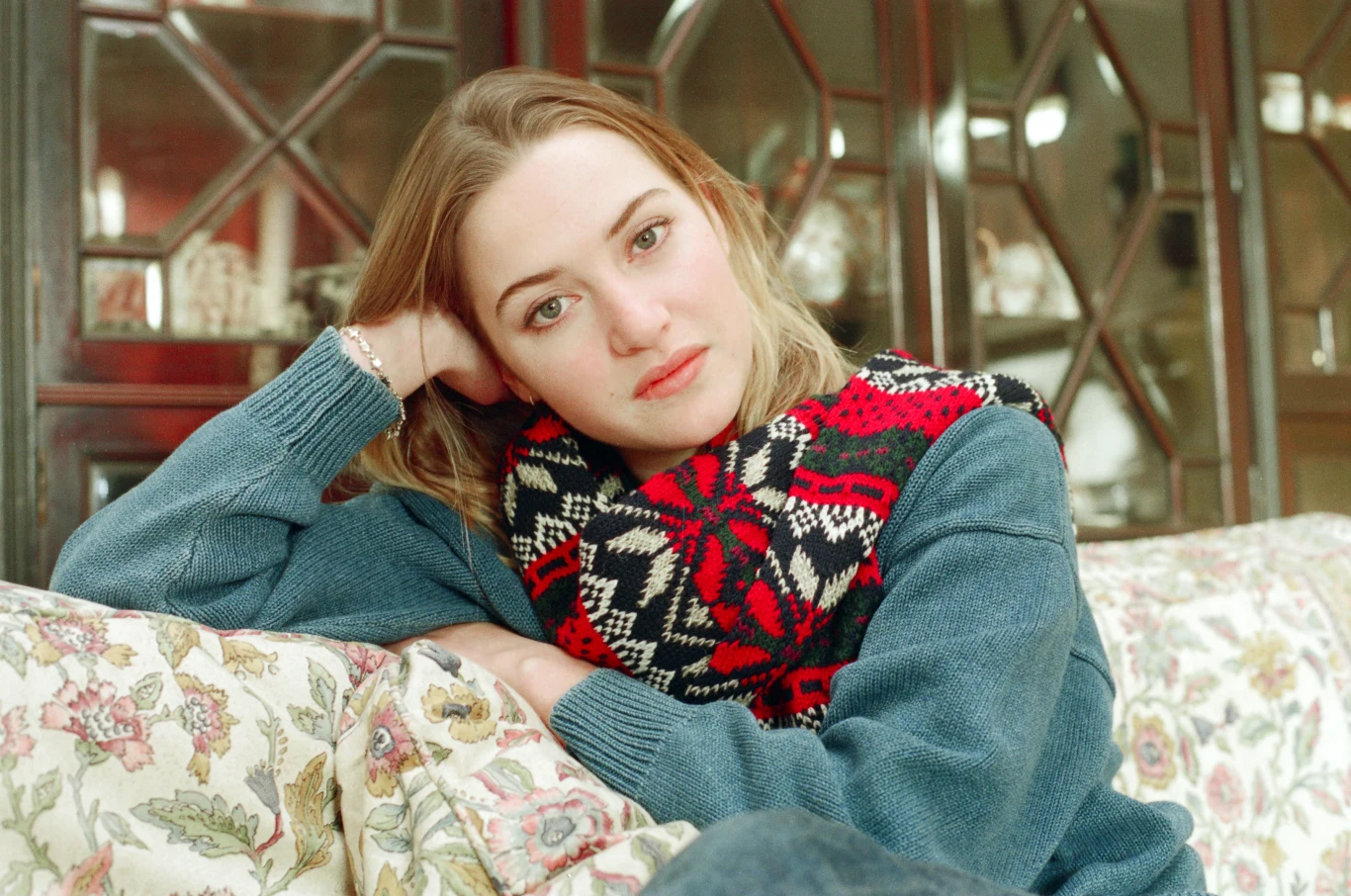 Młoda kobieta z jasnymi włosami siedzi na wzorzystej kanapie, opierając głowę na dłoni, ubrana w niebieski sweter i kolorowy szalik w czerwono-czarny wzór, patrzy spokojnie w stronę aparatu.