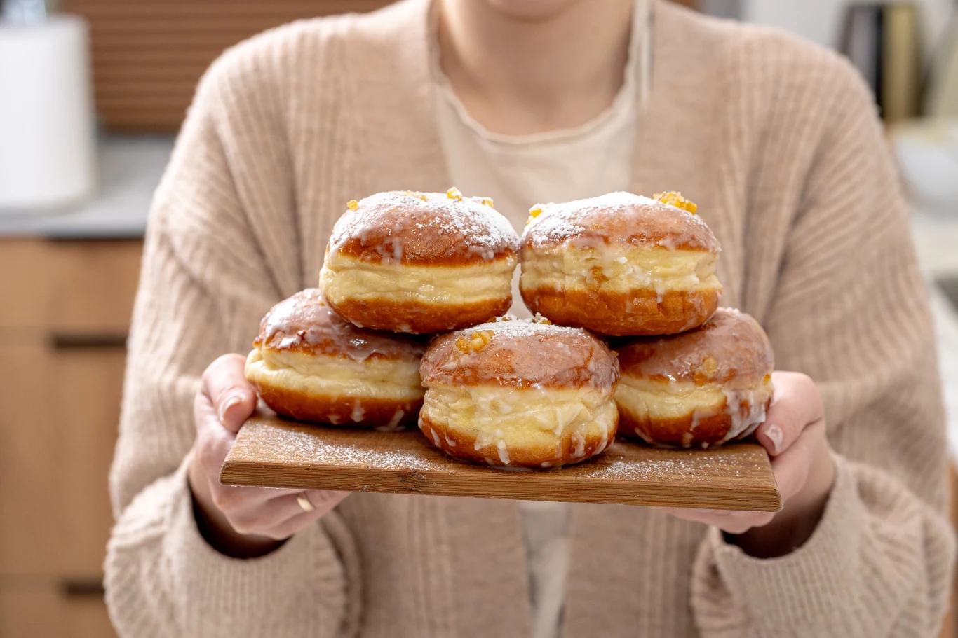 Stos pączków z lukrem i kandyzowaną skórką pomarańczową na drewnianej desce, trzymanej przez osobę ubraną w beżowy sweter.