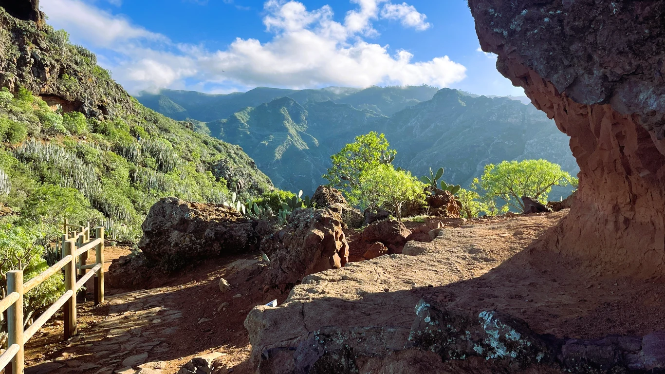 Górska ścieżka prowadząca przez kamienisty teren, otoczona zieloną roślinnością i drewnianym ogrodzeniem, w tle wysokie góry oświetlone promieniami słońca oraz niebieskie niebo z białymi chmurami.