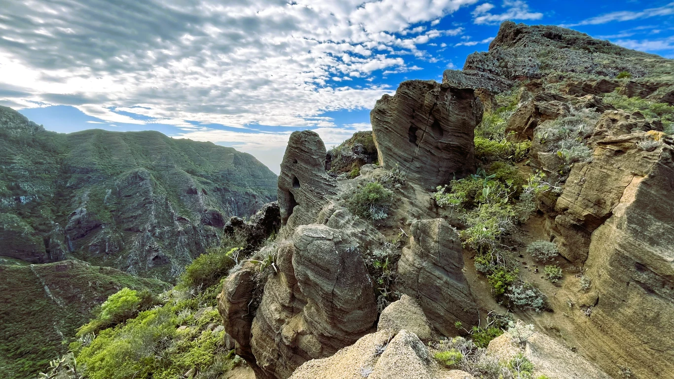 Skały o nieregularnych kształtach porośnięte roślinnością, w tle rozciągające się zielone wzgórza i dramatycznie ukształtowane chmury na niebieskim niebie.