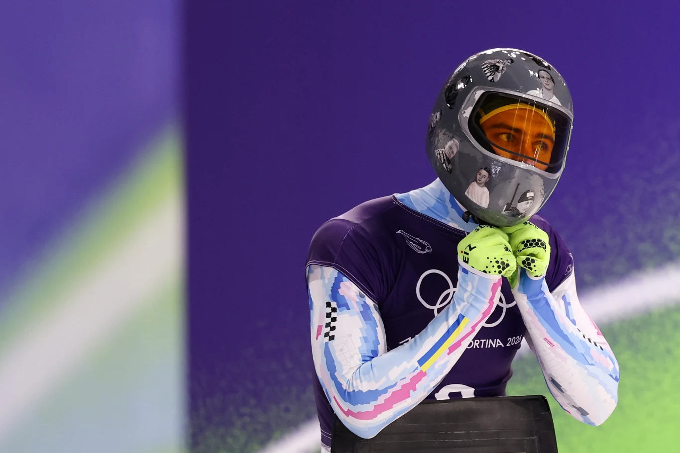 Zawodnik ubrany w sportowy kombinezon oraz kask z przyciemnianą szybą, przygotowujący się do startu na zawodach zimowych, w tle widoczne elementy toru oraz charakterystyczne barwy olimpijskie.