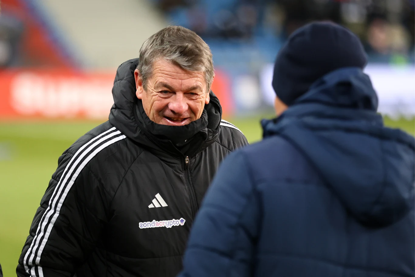 Mężczyzna w sportowej kurtce rozmawia z inną osobą w kurtce z kapturem, znajdują się na stadionie piłkarskim, w tle rozmyte trybuny.