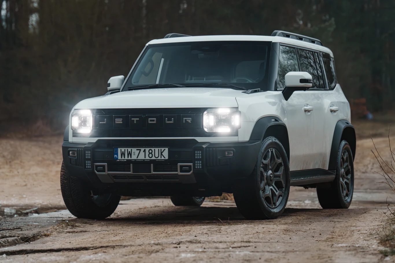 Biały samochód terenowy Jetour T2 z włączonymi światłami przednimi stoi na leśnej drodze, otoczony drzewami w tle, pojazd umieszczony centralnie, widoczna tablica rejestracyjna i wyraziste linie nadwozia.