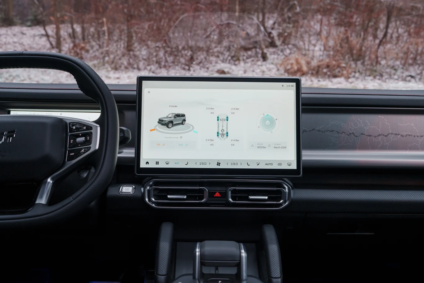 Wnętrze samochodu z centralnie umieszczonym dużym ekranem dotykowym, na którym wyświetlane są ustawienia i informacje dotyczące pojazdu, kierownica po lewej stronie, przez szybę widoczne zaśnieżone drzewa.