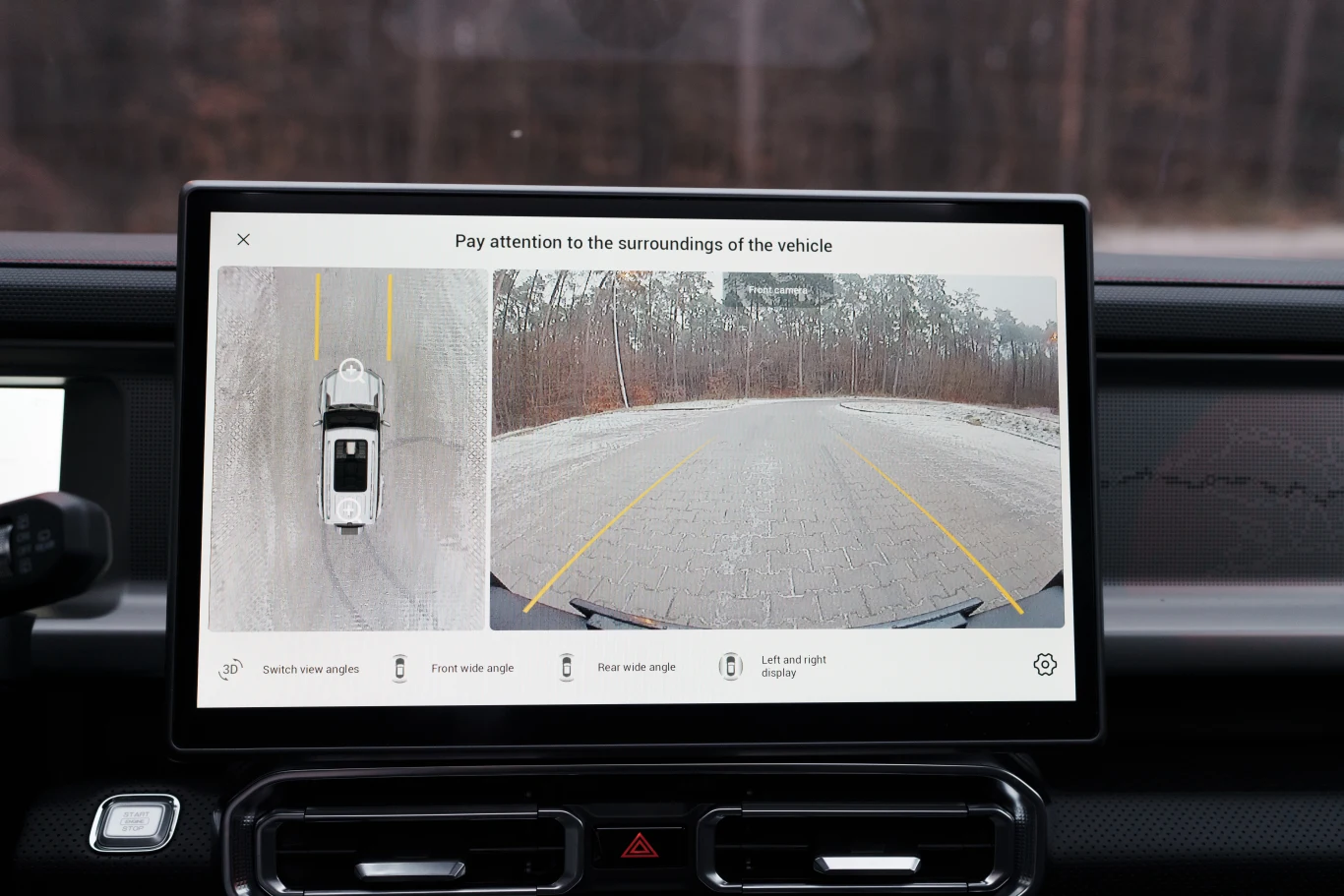 Panel wyświetlający obraz z kamery cofania oraz widok z góry na samochód, żółte linie pomocnicze ułatwiają manewrowanie, w tle widoczne drzewa i droga pokryta śniegiem.