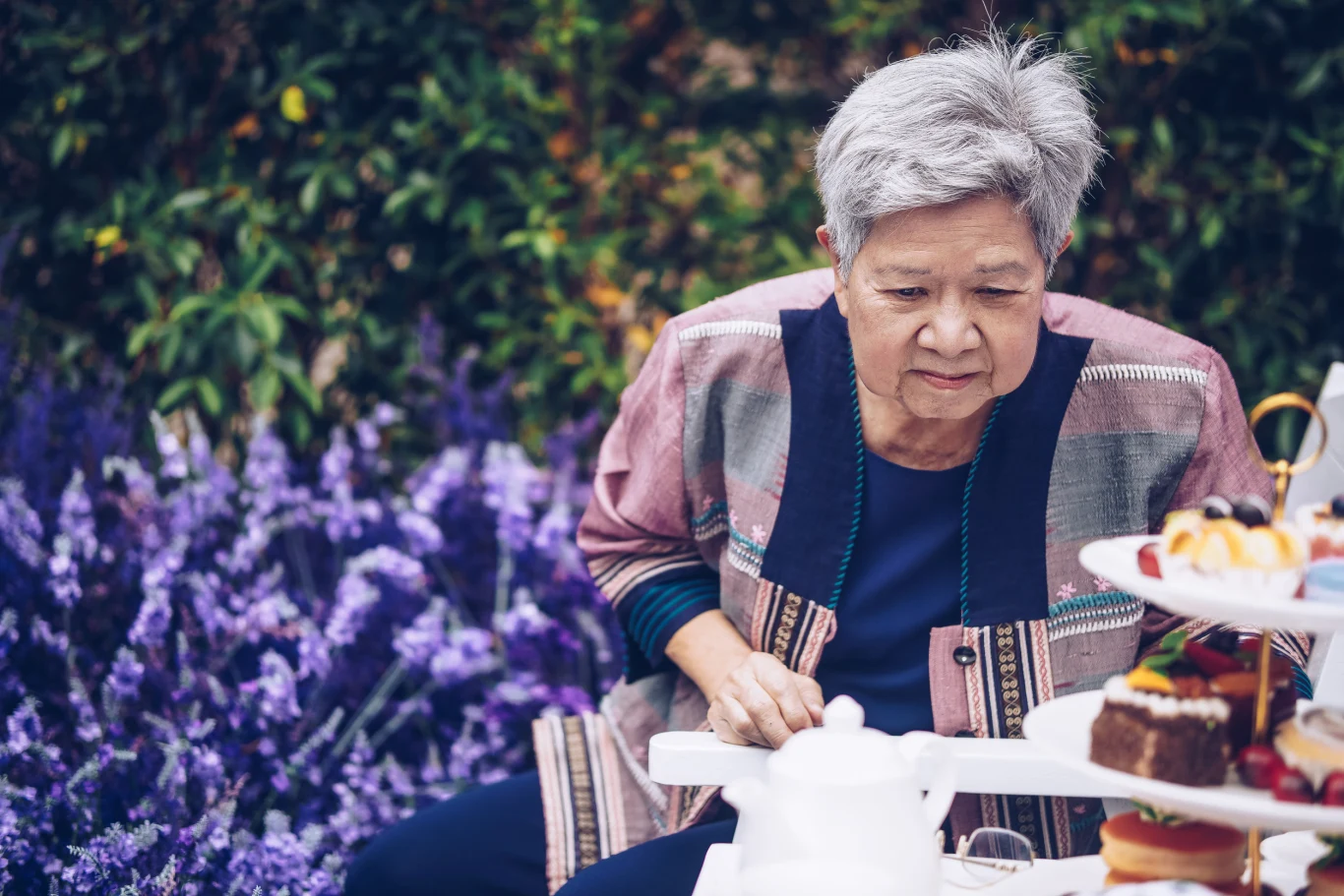 Starsza kobieta siedzi przy stole z przekąskami i ciastkami, w otoczeniu fioletowych kwiatów w ogrodzie.