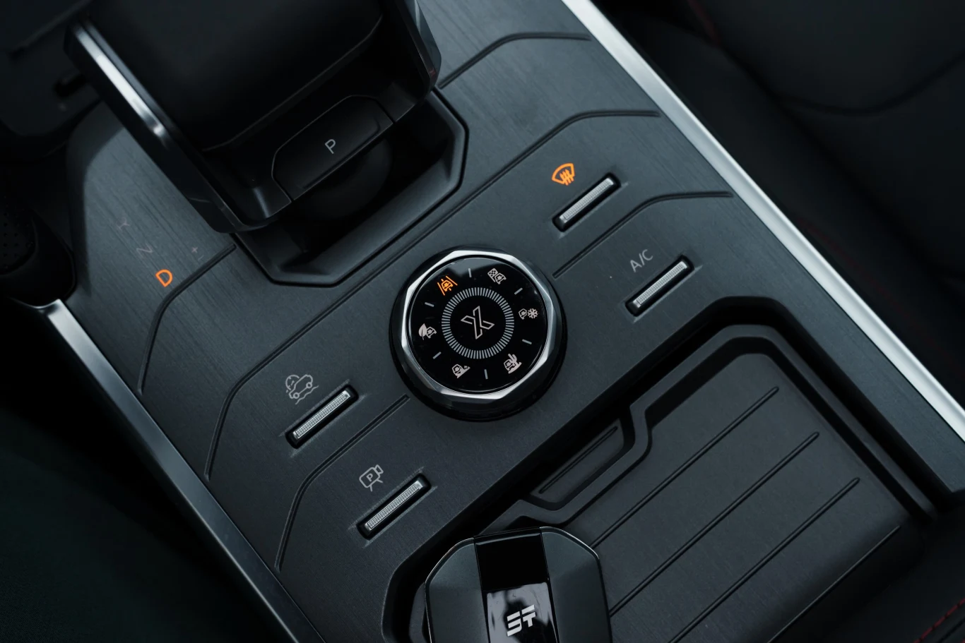 Panel środkowy samochodu z pokrętłem sterującym trybami jazdy, wyraźnie widocznymi przyciskami do sterowania klimatyzacją i ogrzewaniem szyb oraz schowkiem na drobne przedmioty.