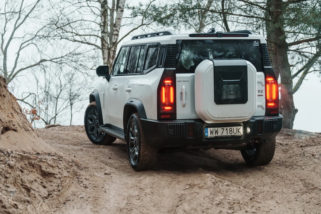 Samochód terenowy w kolorze białym stoi na piaszczystej, leśnej drodze w otoczeniu drzew, widok od tyłu, widoczne koło zapasowe na tylnej klapie oraz tablica rejestracyjna.