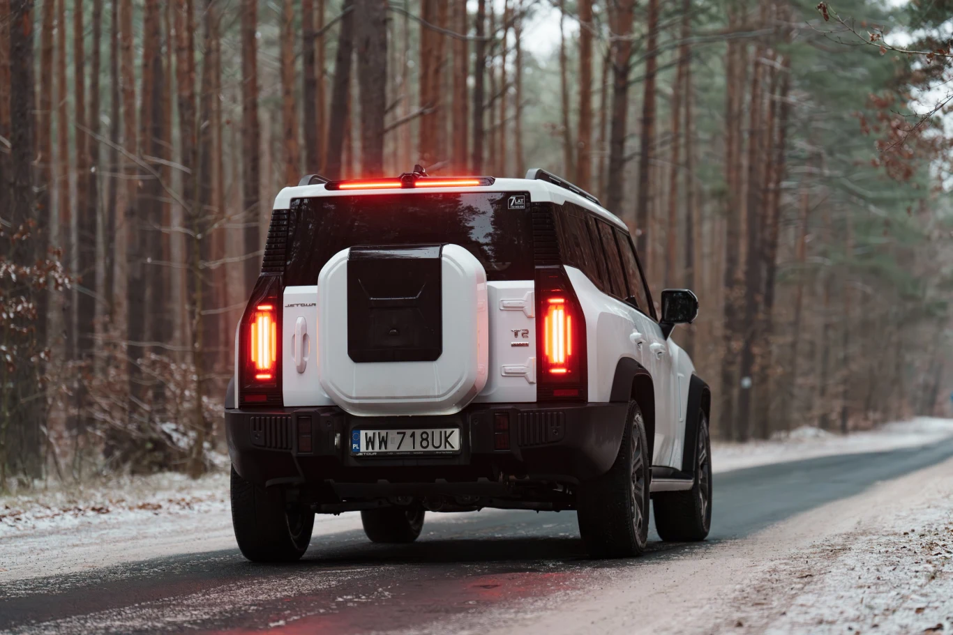 Biały samochód terenowy jedzie leśną, ośnieżoną drogą pośród wysokich drzew. Tylne światła auta są włączone, a droga częściowo pokryta śniegiem.