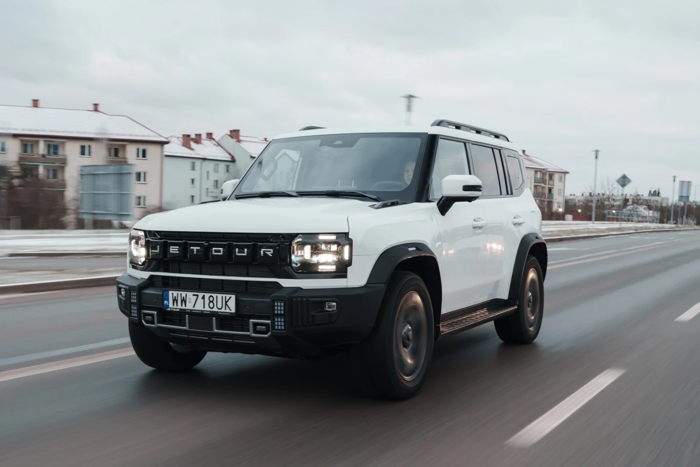 Biały SUV z czarnymi detalami jadący po szerokiej, miejskiej drodze, w tle widać zabudowania wielorodzinne i lekko zachmurzone niebo.