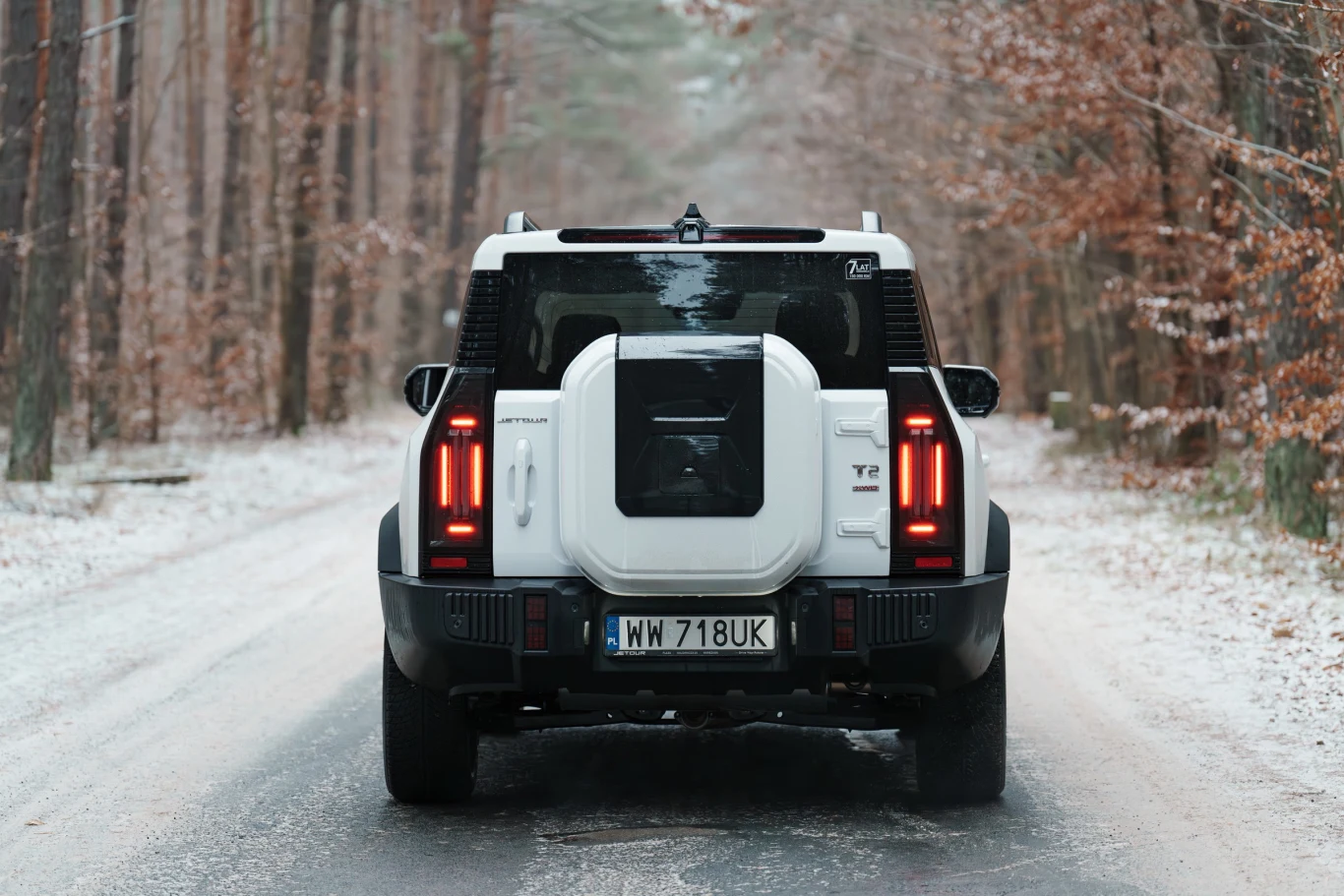 Terenowy samochód marki Land Rover Defender w kolorze białym, widziany od tyłu, stoi na zaśnieżonej leśnej drodze otoczonej drzewami bez liści.