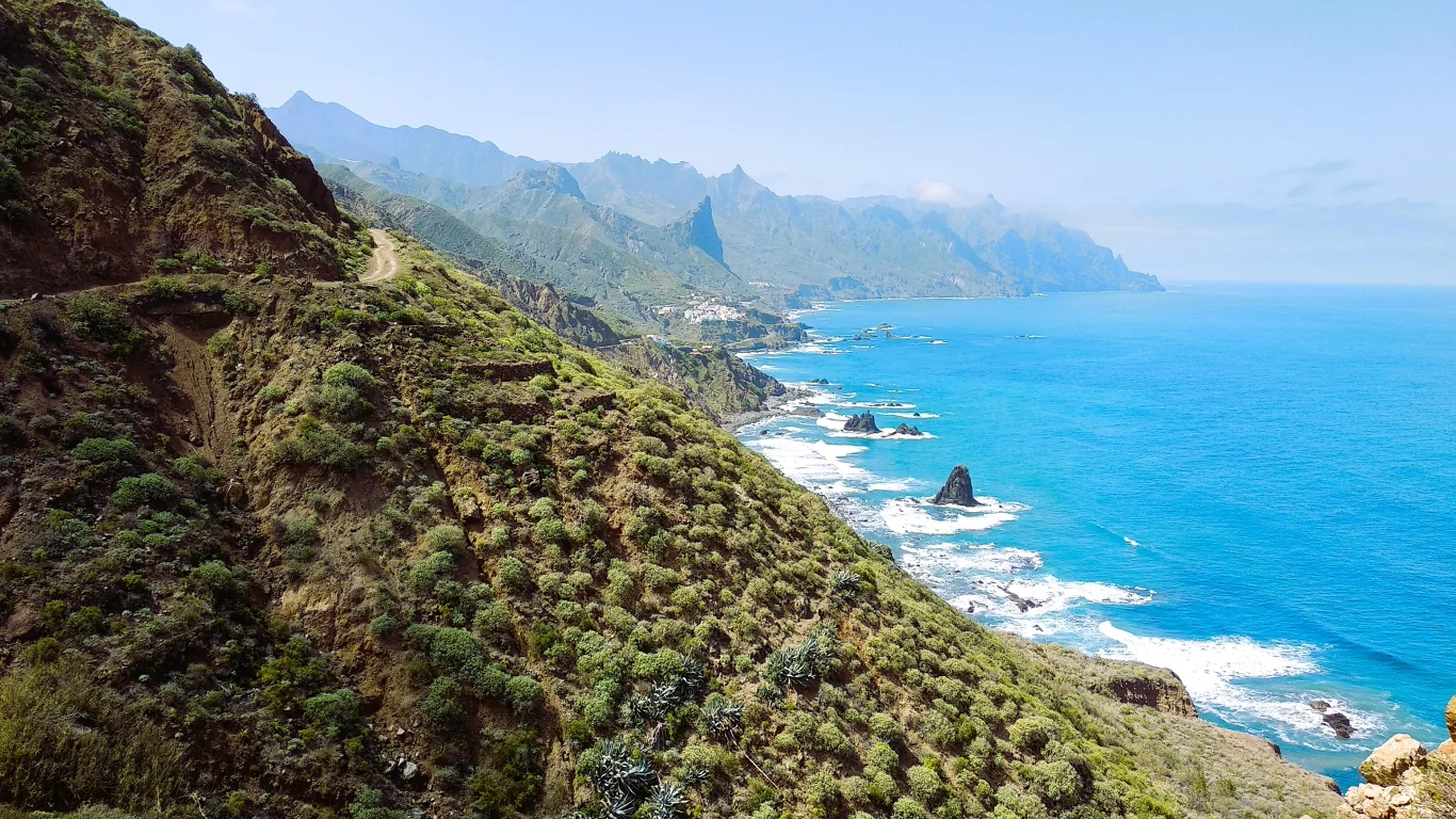 Strome, zielone zbocza gór schodzące do lazurowego oceanu, skaliste formacje wystające z wody oraz rozległa panorama pasma górskiego na dalszym planie, podkreślona wyraźnym niebieskim niebem