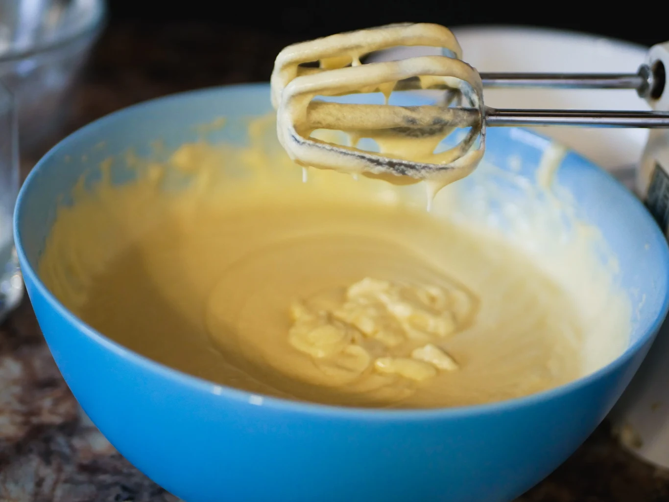 Miska z jasnożółtym ciastem w trakcie miksowania, metalowe końcówki miksera pokryte kremową masą unoszą się nad ciastem.