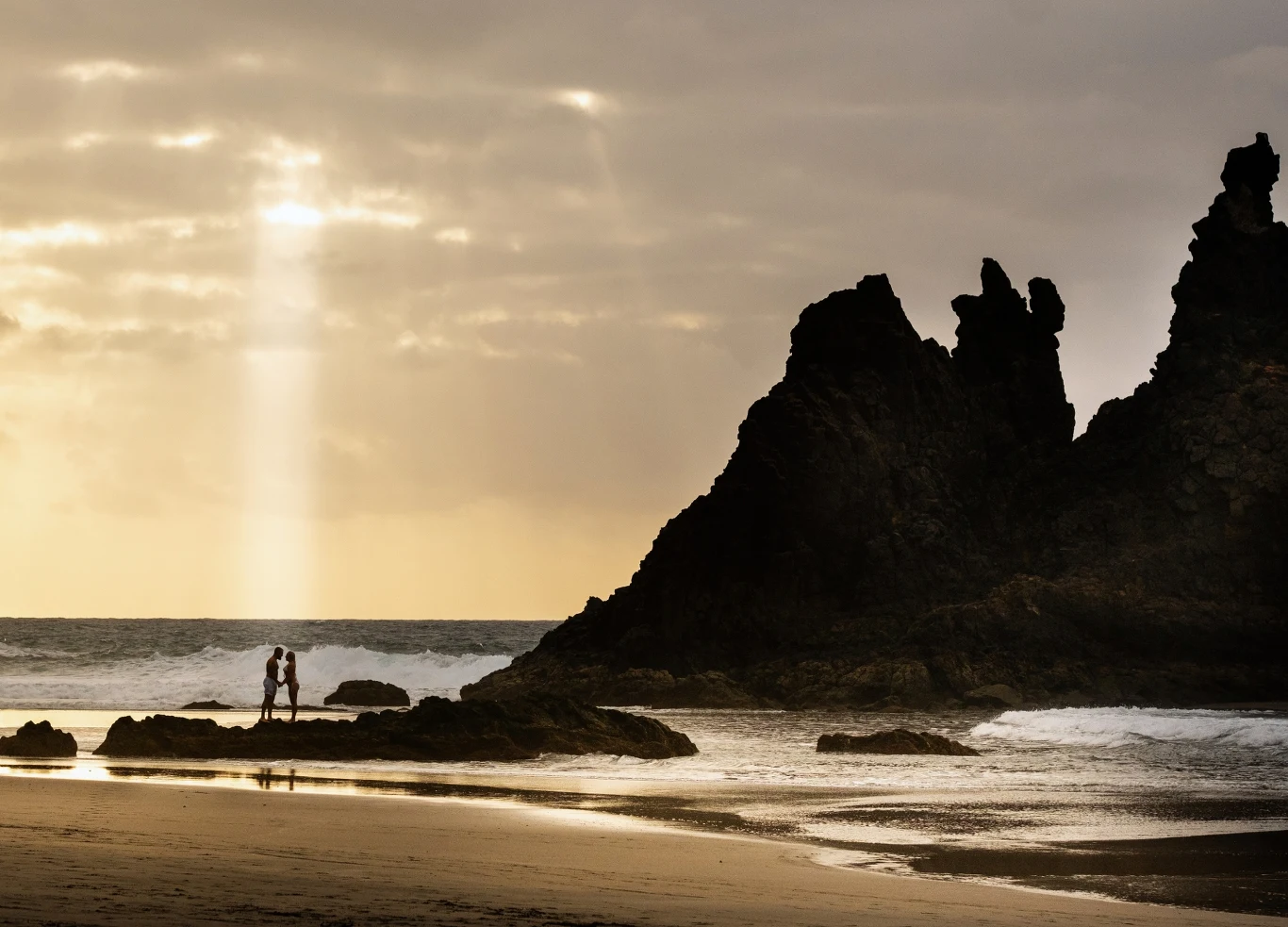 Plaża o zachodzie słońca z ciemnymi, strzelistymi formacjami skalnymi po prawej stronie oraz promieniem światła przebijającym się przez chmury. Dwie osoby stoją na brzegu, blisko wody, otoczone spokojną, naturalną scenerią.