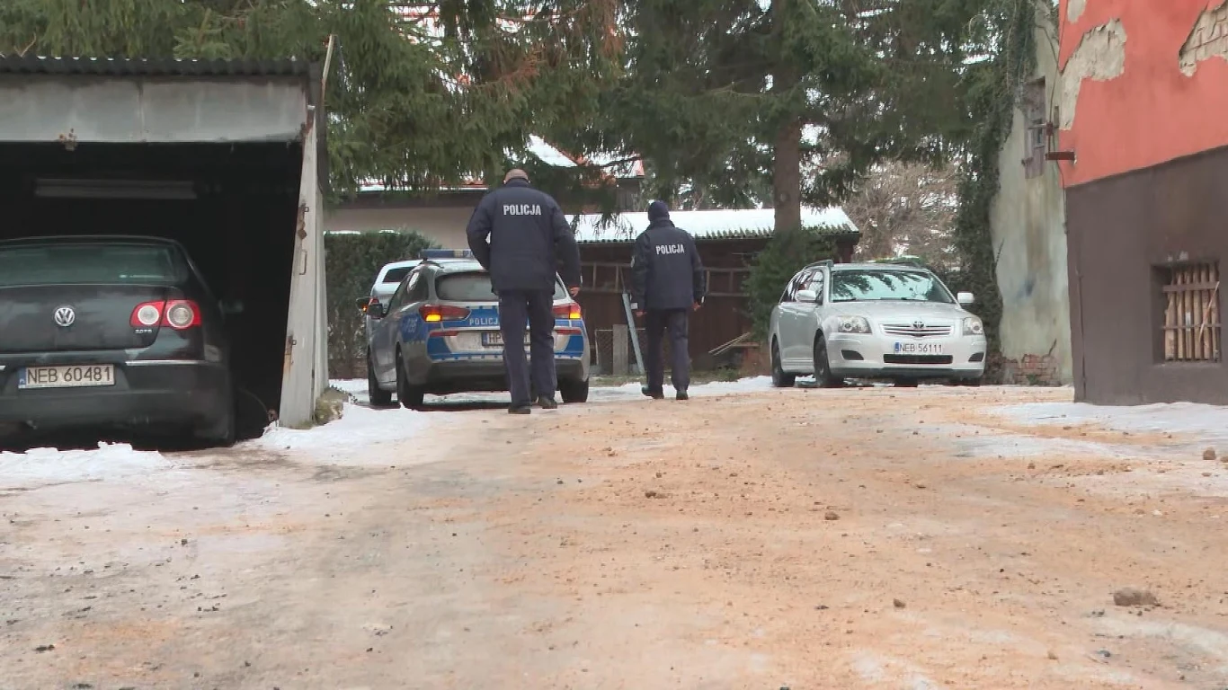 Dymisja po aferze na komendzie w Tolkmicku Dwóch policjantów idzie po zaśnieżonym podwórku w kierunku zaparkowanych samochodów, obok garażu, w zimowym otoczeniu wśród drzew.