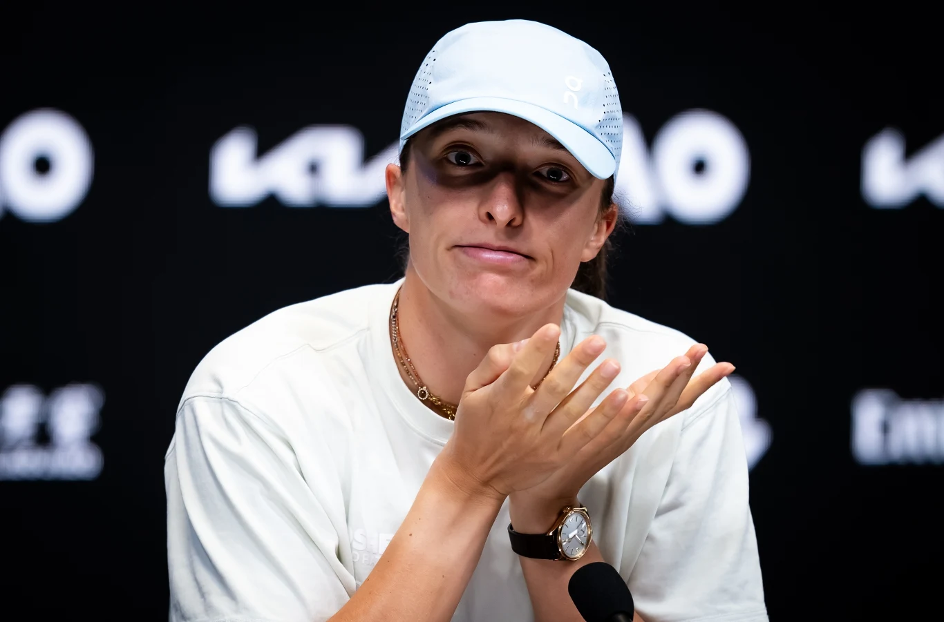 Iga Świątek Osoba w jasnej czapce i białej koszulce siedzi przy mikrofonie na tle czarnego ekranu z białymi napisami, wykonuje gest rękami i patrzy prosto do obiektywu.