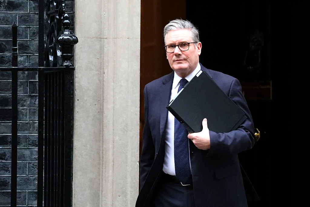 Premier Wielkiej Brytanii Keir Starmer nie zamierza podawać się do dymisji Mężczyzna w garniturze i okularach wychodzi z budynku, trzymając w ręce czarną teczkę. Tło stanowią drzwi wejściowe i fragment ceglanej ściany.