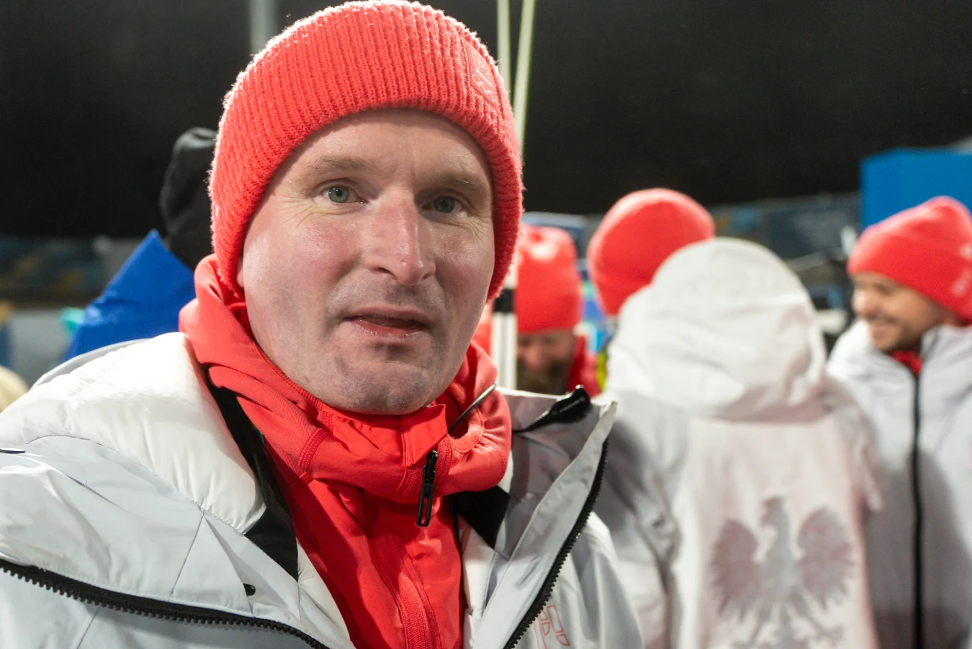 Mężczyzna ubrany w białą kurtkę i czerwoną czapkę stoi na pierwszym planie, w tle kilka osób również w zimowych ubraniach i czerwonych czapkach, fragment kurtki jednej osoby ozdobiony godłem Polski.