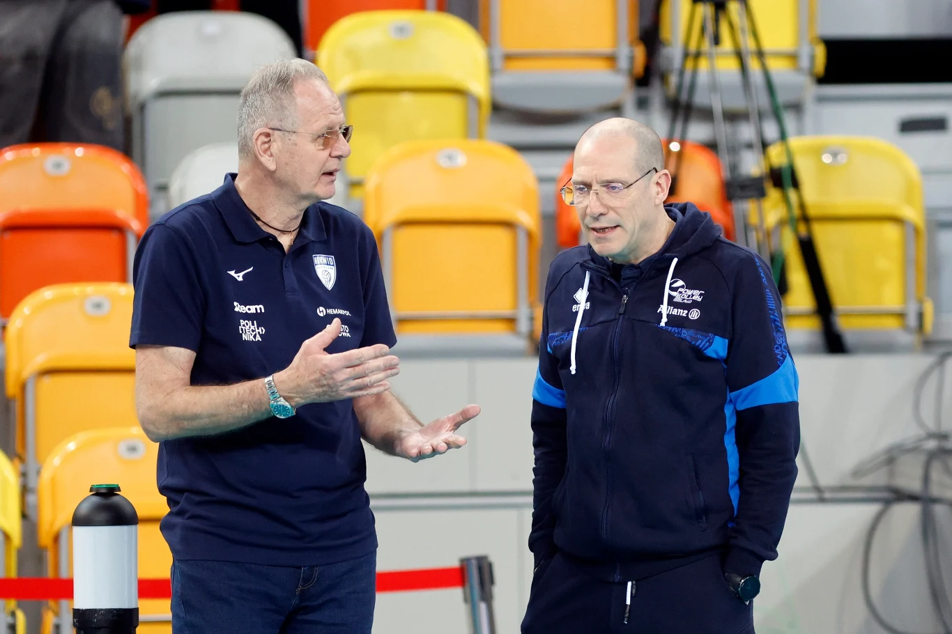 Trenerzy drużyn z Częstochowy i Mediolanu, Ljubomir Travica (z lewej) i Roberto Piazza Dwóch mężczyzn w sportowych ubraniach prowadzi rozmowę na tle żółtych i pomarańczowych trybun na hali sportowej. Jeden z nich gestykuluje rękami, drugi słucha z zainteresowaniem.