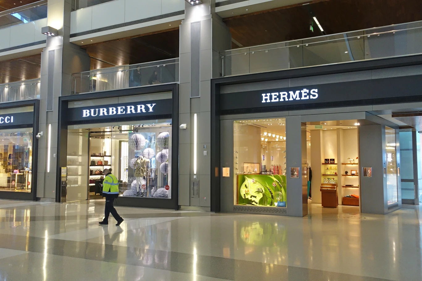 Komisja Europejska zakazała niszczenia nowej odzieży i nowego obuwia. Nowe regulacje mogą uderzyć w luksusowe marki Witryny sklepów luksusowych Burberry i Hermès w nowoczesnym pasażu handlowym z osobą w żółtej kamizelce odblaskowej przechodzącą przed wejściem, wnętrza sklepów oświetlone, prezentujące asortyment.