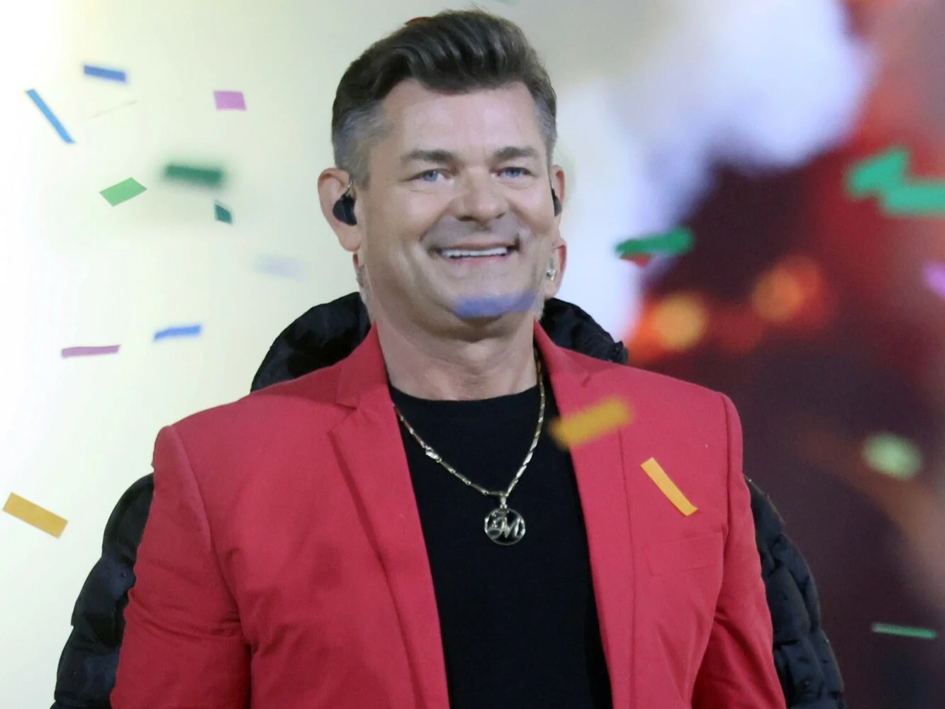Mężczyzna w czerwonej marynarce z krótkimi włosami i łańcuszkiem na szyi, stojący na tle kolorowych konfetti oraz rozmytego tła z jasnym światłem.