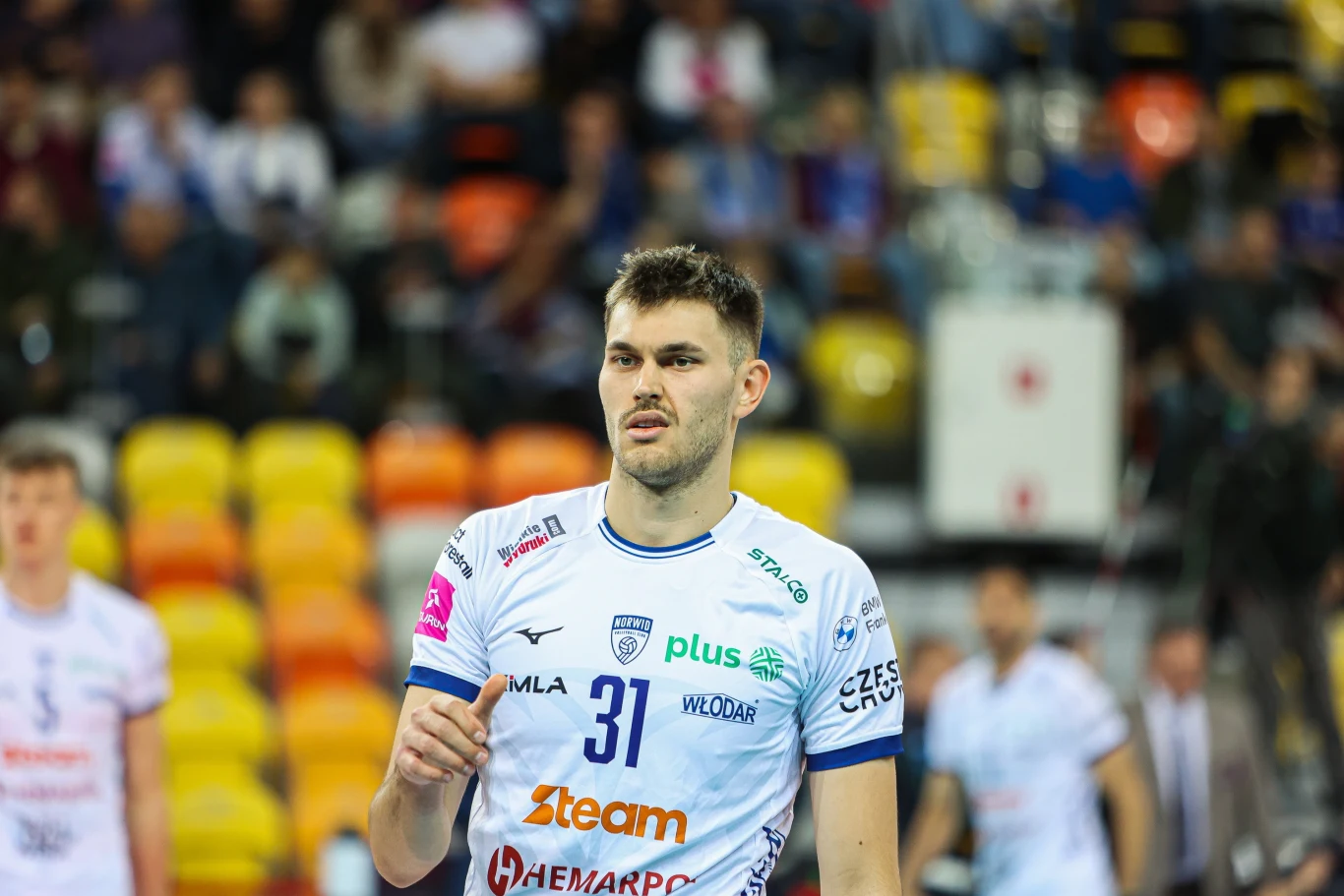 Sebastian Adamczyk, środkowy Steam Hemarpol Politechniki Częstochowa Siatkarz w białej koszulce z numerem 31, przebywający na hali sportowej podczas meczu, w tle widoczni inni zawodnicy oraz rozmyte trybuny z publicznością.