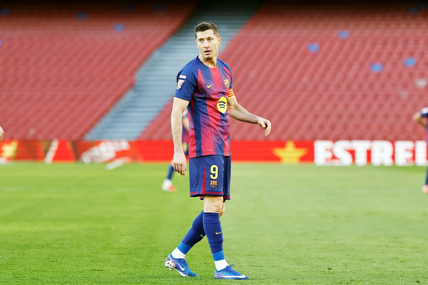 Piłkarz w stroju meczowym drużyny FC Barcelona stoi na murawie stadionu, w tle puste trybuny.