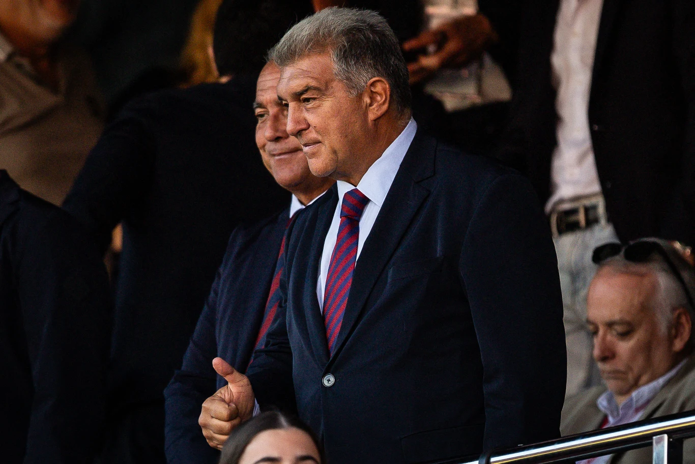 Joan Laporta Mężczyźni w garniturach i krawatach stoją w tłumie, jeden z nich wykonuje gest kciuka w górę i uśmiecha się, w tle widoczne inne osoby.