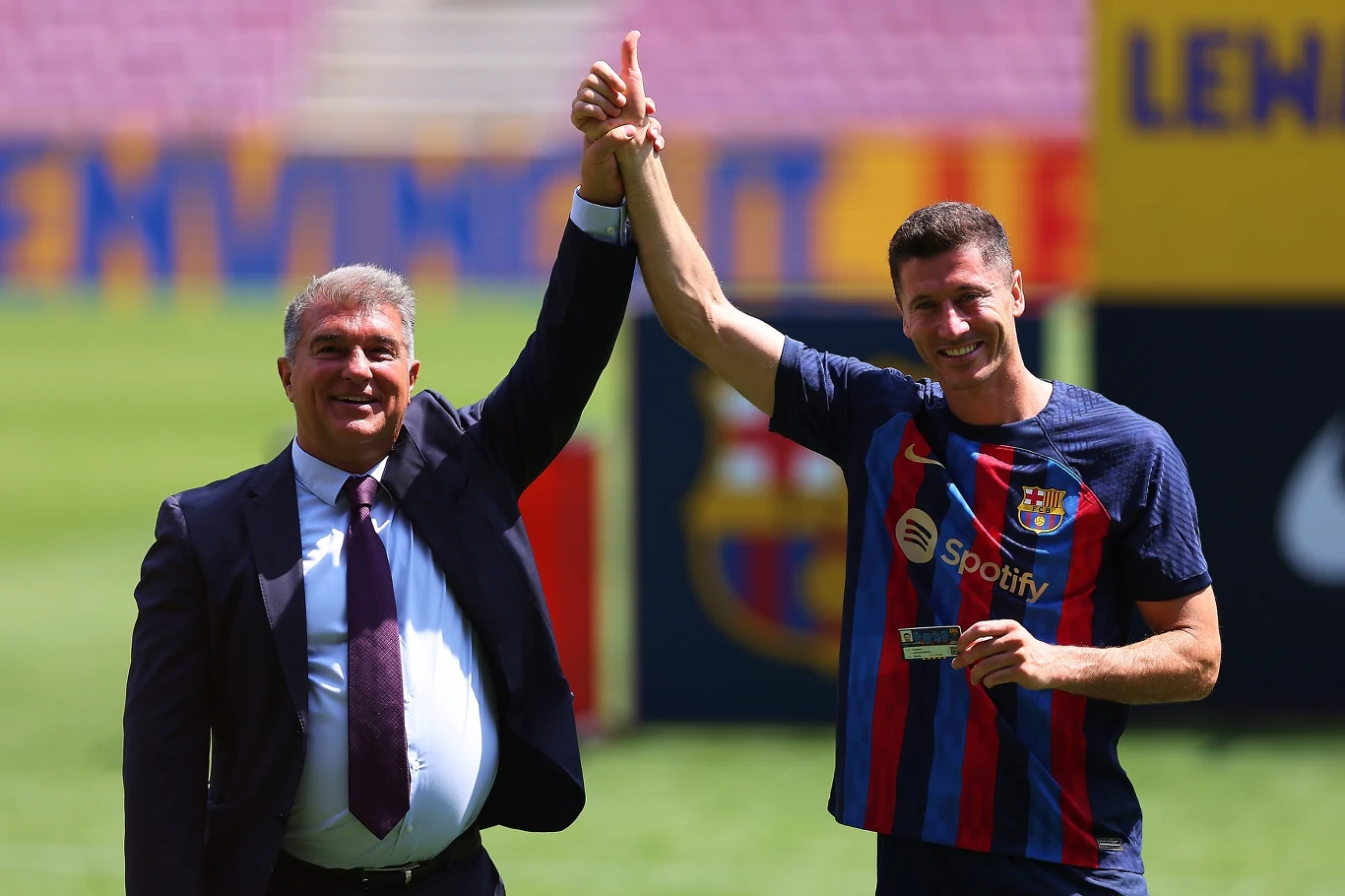 Dwóch mężczyzn na tle stadionu; jeden z nich w garniturze, drugi w stroju piłkarskim FC Barcelony podnosi rękę drugiego do góry, obaj uśmiechnięci. W tle elementy stadionu i emblemat klubu.