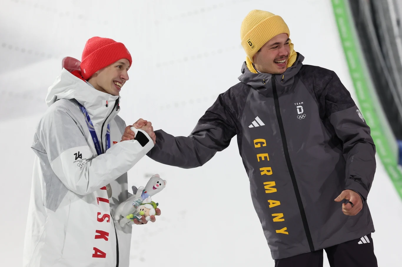 Dwóch sportowców w zimowych strojach narodowych Polski i Niemiec okazuje wzajemny szacunek, ściskając sobie dłonie podczas ceremonii, w tle widoczne elementy skoczni narciarskiej.