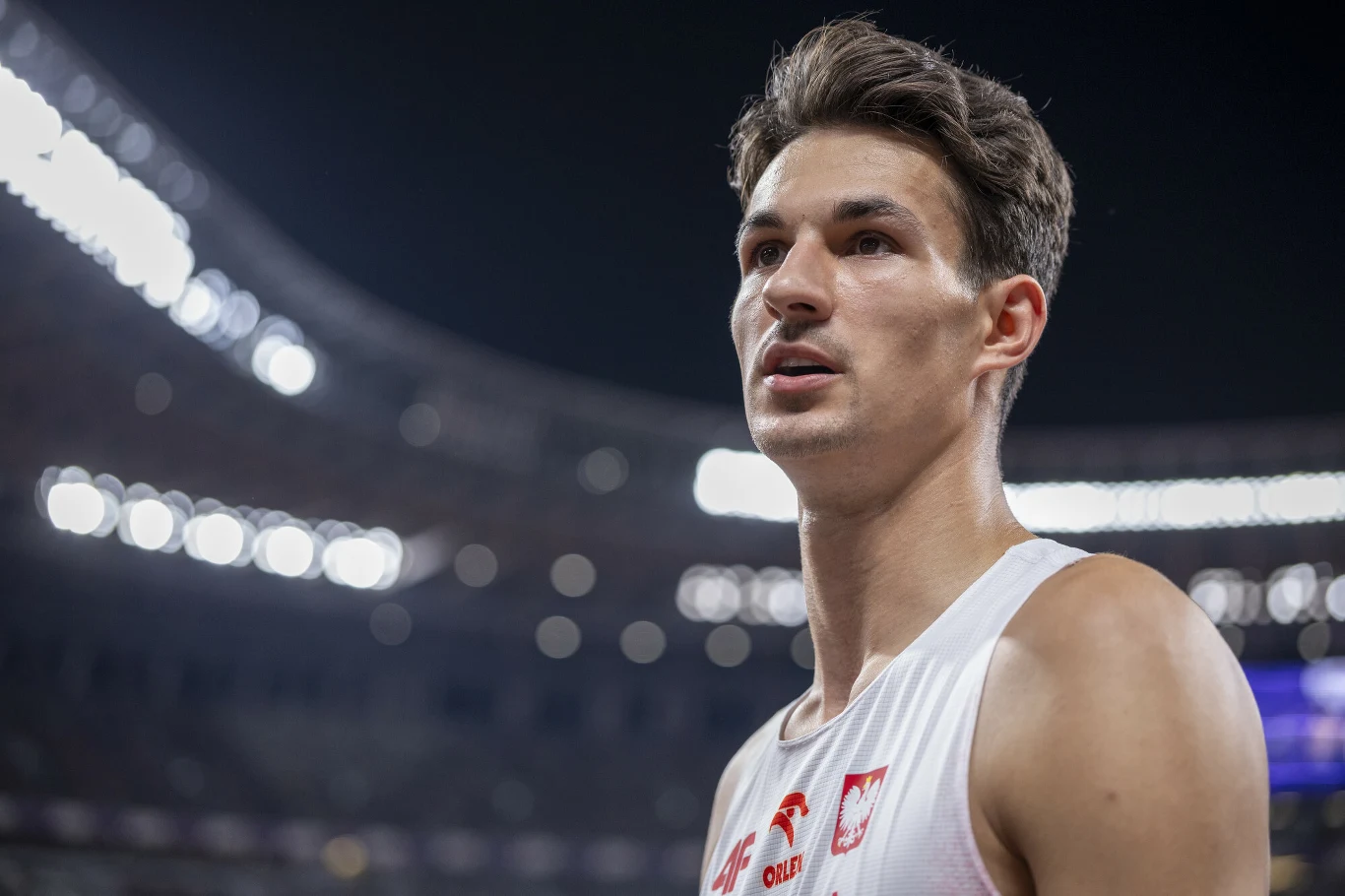 Młody sportowiec w stroju reprezentacji Polski na tle stadionu nocą, wyraźnie skupiony, światła i rozmyte elementy w tle podkreślają atmosferę sportowej rywalizacji.